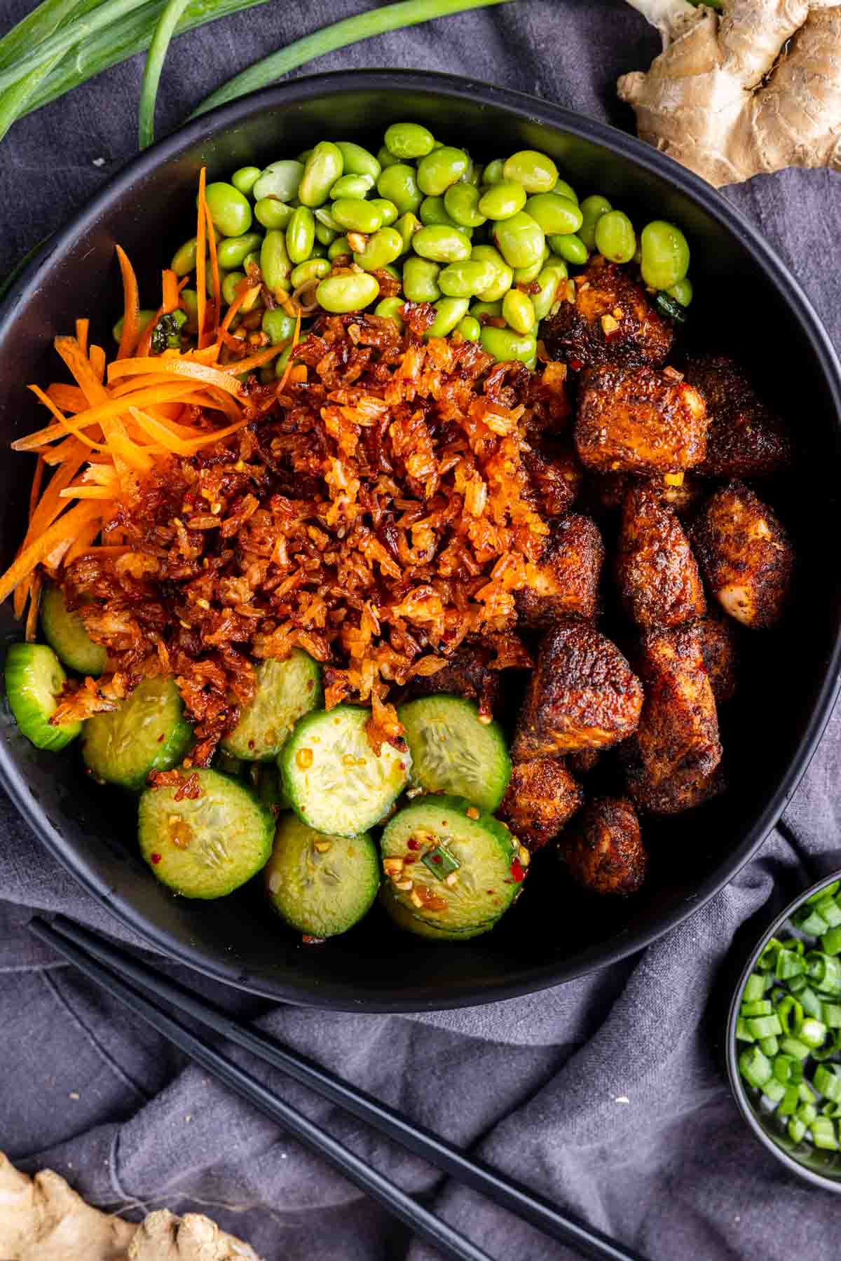 A bowl containing cooked rice, cubes of seasoned salmon, sliced cucumber, shredded carrots, and edamame, arranged in sections—like a meal prep salmon bowl. Black chopsticks rest beside the bowl. Try with chili crisp dressing for extra flavor!.