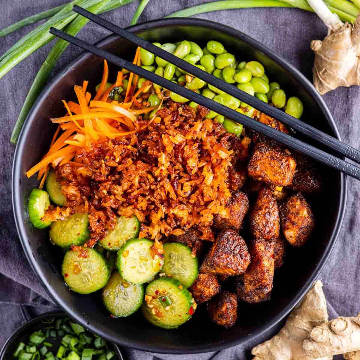 A black bowl filled with rice, seasoned tofu, sliced cucumbers, shredded carrots, edamame, and chopsticks, surrounded by scallions and ginger on a gray cloth—perfect inspiration for your next air fryer salmon bites recipe.