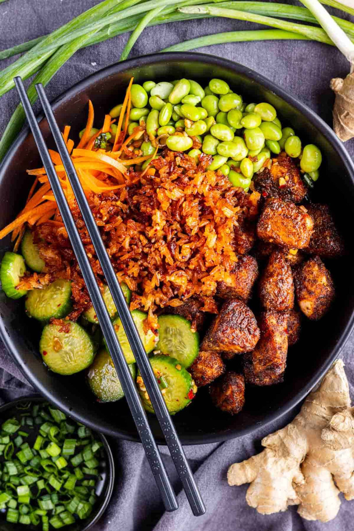 A black bowl filled with cubed seasoned chicken, sliced cucumbers, shredded carrots, edamame, crispy onions, and black chopsticks, with ginger and green onions on the side.