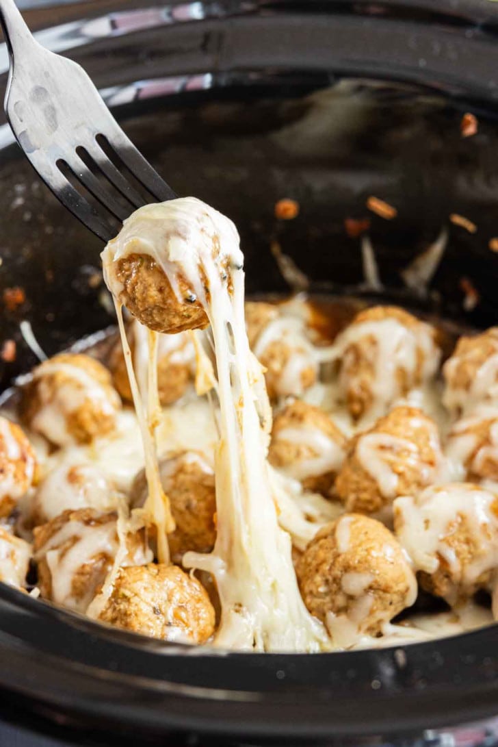 A fork lifts a cheese-covered meatball from a slow cooker filled with melted cheese and more meatballs—these crockpot French onion meatballs make an easy appetizer everyone will love.