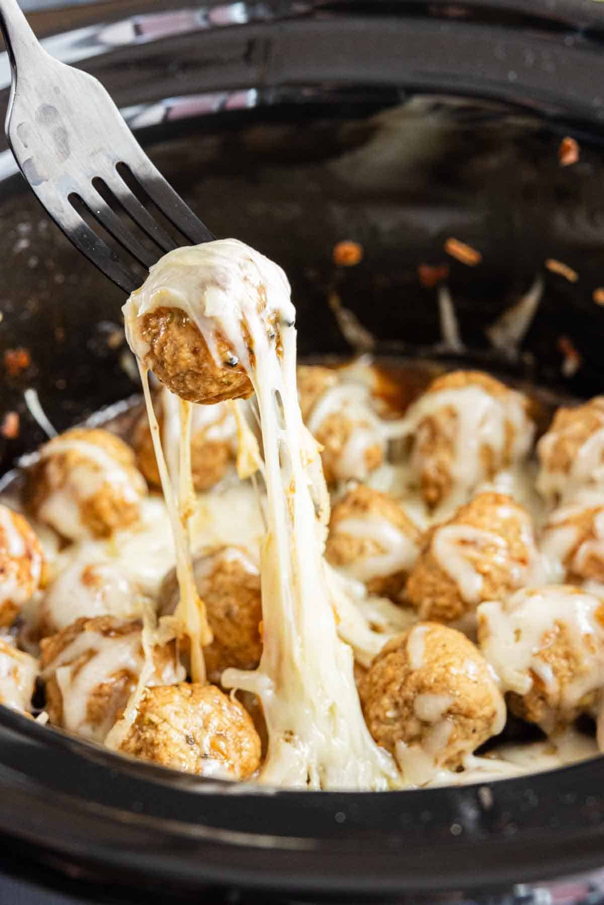 A fork lifts a cheese-covered meatball from a slow cooker filled with melted cheese and more meatballs—these crockpot French onion meatballs make an easy appetizer everyone will love.