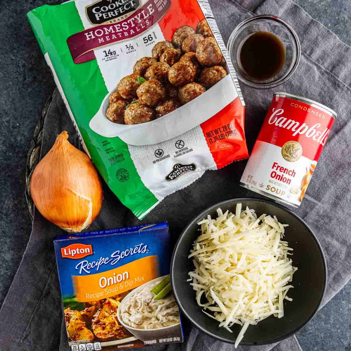 Frozen meatballs, a can of French onion soup, a yellow onion, onion soup mix, shredded cheese, and a small bowl of sauce arranged on a gray cloth—perfect for an easy appetizer meatballs dish or your next French onion meatballs recipe.