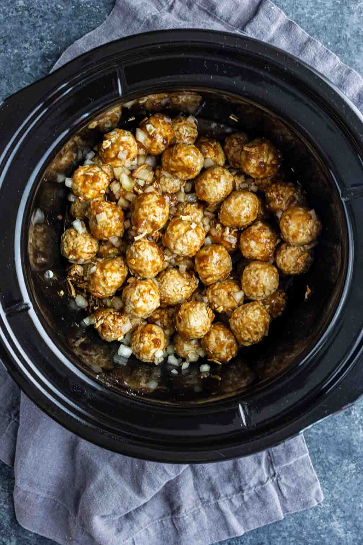 Uncooked frozen meatballs mixed with chopped onions and French onion soup mix inside a black slow cooker, placed on a gray cloth on a dark countertop.
