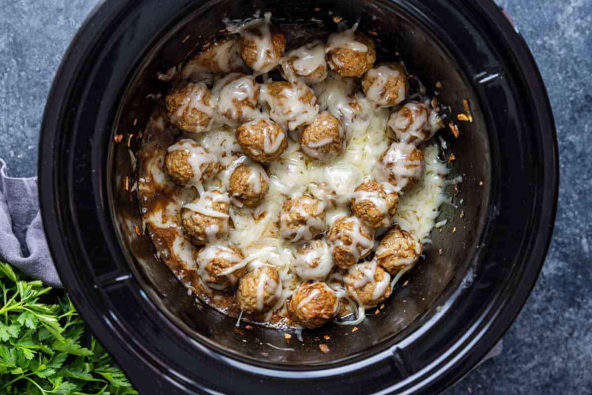 Cooked crockpot French onion meatballs topped with melted cheese inside a black slow cooker, with some parsley visible on the side—an easy appetizer meatballs recipe perfect for gatherings.