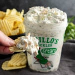 A hand holds a ridged potato chip dipped in creamy Viral Pickle Dip, with a container of pickle dip and a pile of potato chips in the background.