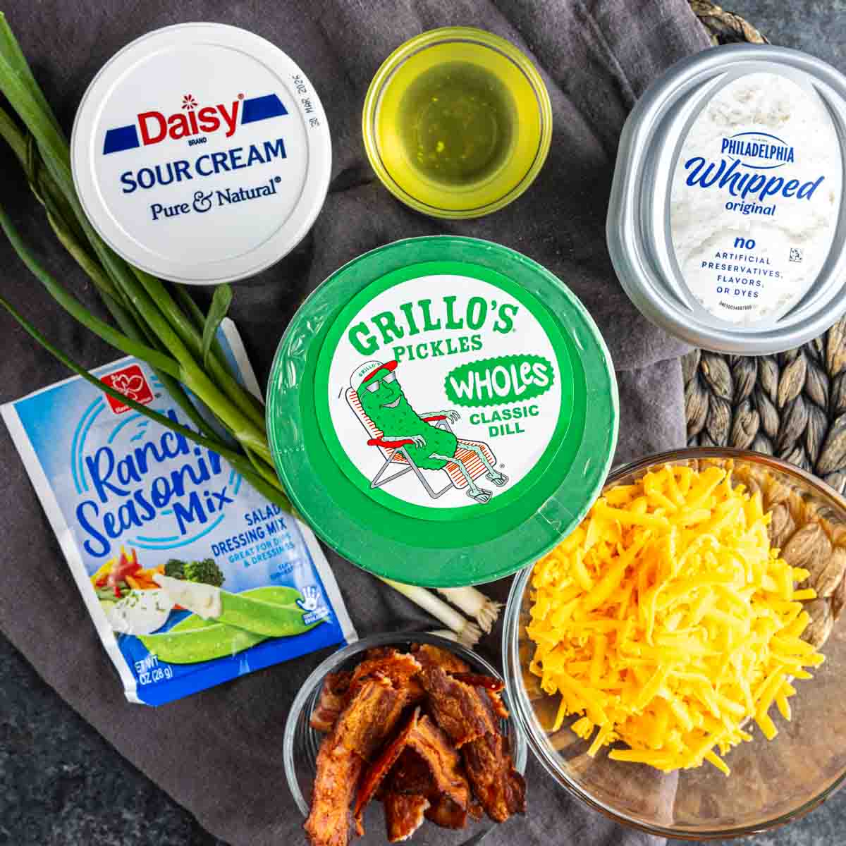 Assorted ingredients displayed on a table, ready to make the viral pickle dip: sour cream, whipped cream cheese, ranch seasoning mix, cheddar cheese, cooked bacon, green onions, pickles, and a small bowl of oil.