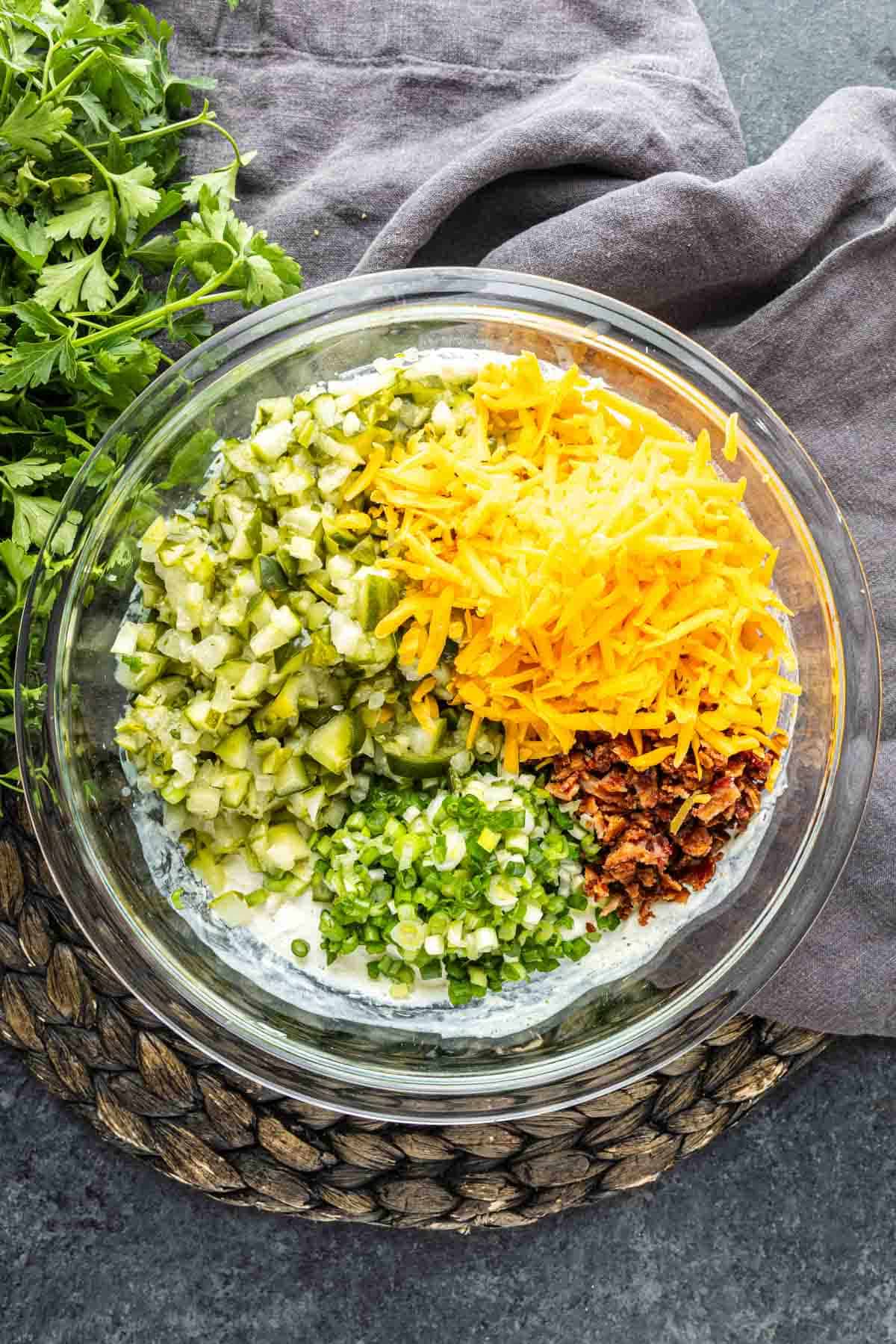A glass bowl with chopped pickles, shredded cheddar cheese, green onions, and bacon bits on a creamy base—this cold party dip, inspired by the Viral Pickle Dip trend on TikTok, sits next to fresh cilantro on a dark surface.