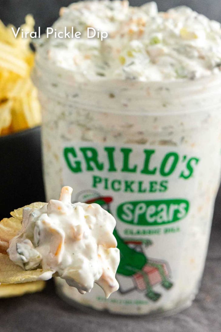 A container of Grillo’s Pickles spears filled with creamy pickle dip, with a chip topped with the dip in the foreground and potato chips in the background. Text reads "Viral Pickle Dip.