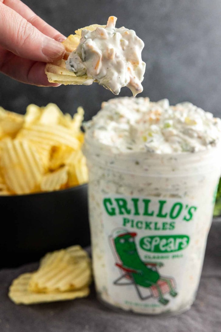 A hand dips a potato chip into a container of homemade vegetable dip in a Grillo’s Pickles jar, with a bowl of ridged potato chips in the background.