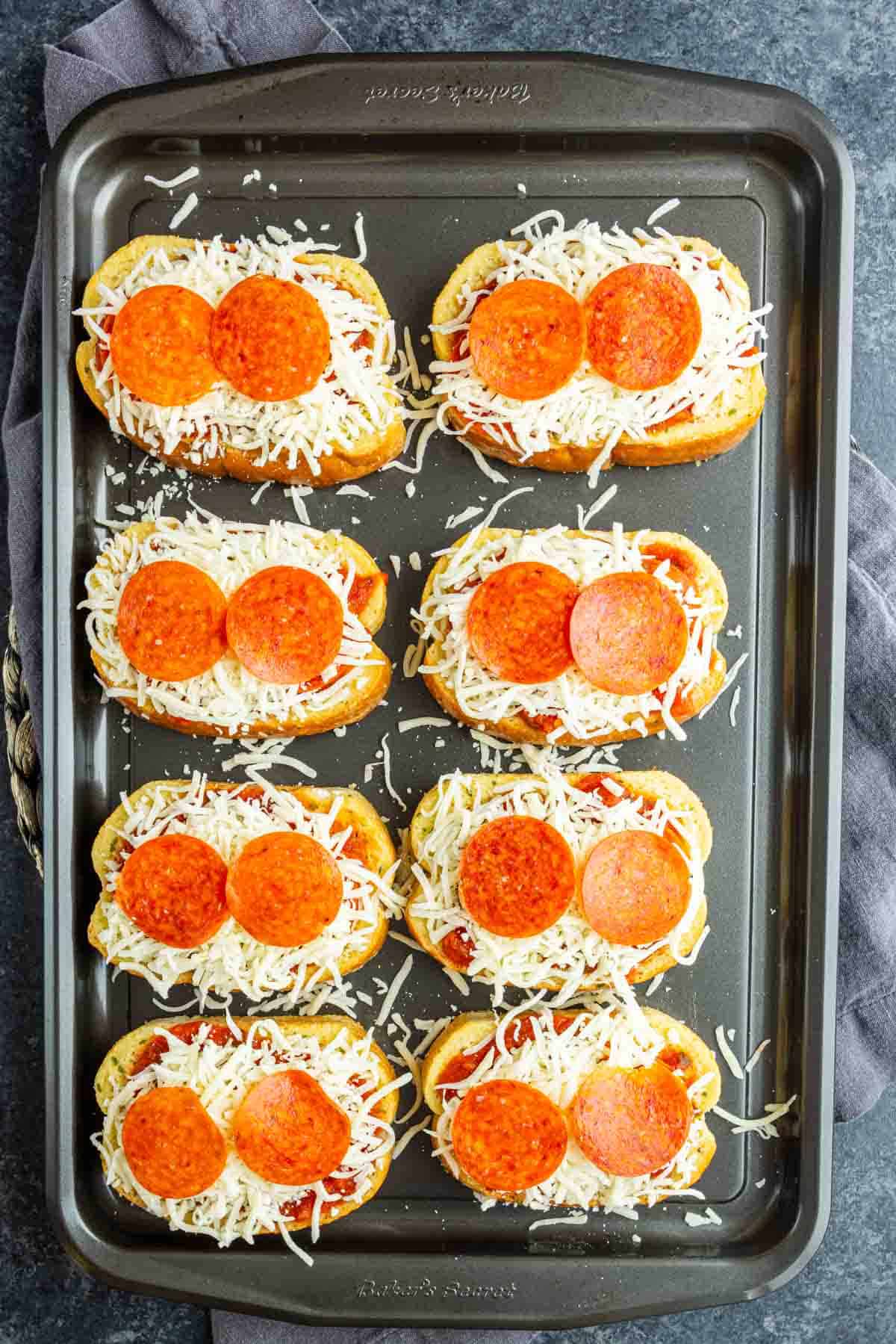 Eight slices of Texas Toast Pizza are topped with shredded cheese and pepperoni, arranged on a baking sheet—an easy pizza toast recipe ready to be baked.