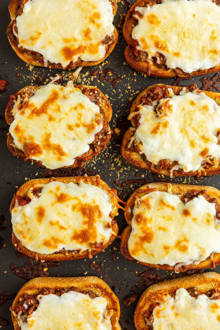Nine pieces of toasted bread topped with meat sauce and melted, browned cheese are arranged on a dark baking sheet, creating an easy Garlic Bread Lasagna twist that's perfect for Texas Toast Lasagna fans.