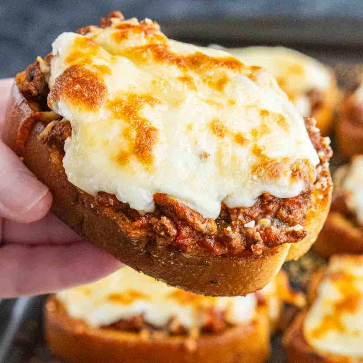 A hand holds a slice of easy garlic bread lasagna—seasoned ground beef, tomato sauce, and melted cheese atop thick bread; more slices are visible in the background.