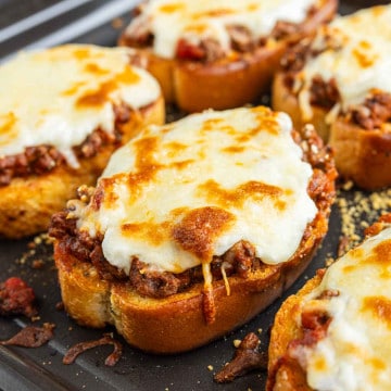 Slices of toasted bread topped with seasoned ground beef, tomato sauce, and melted cheese are arranged on a baking sheet.