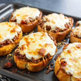 Six slices of Texas Toast Lasagna on a baking tray, each topped with seasoned ground beef, tomato sauce, and melted mozzarella cheese for a delicious twist on classic garlic bread.