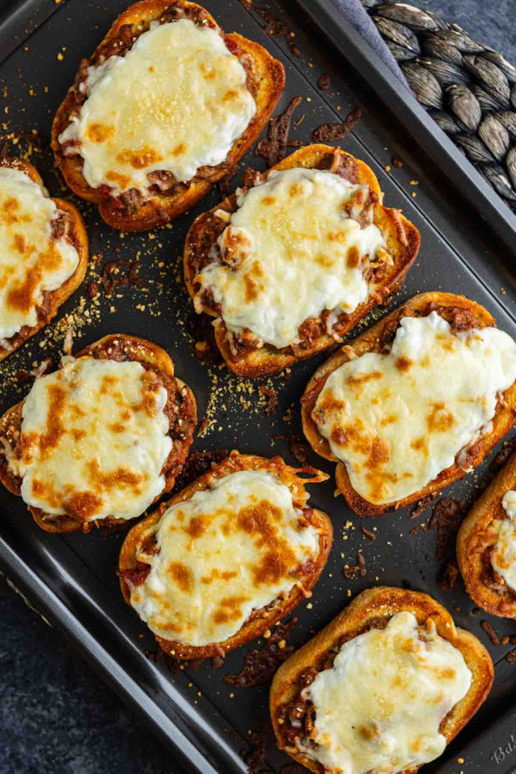 Eight slices of toasted bread topped with meat sauce and melted cheese arranged on a dark baking sheet.
