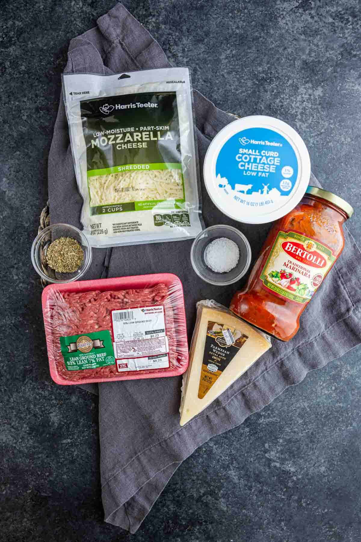Ingredients for an easy garlic bread lasagna are laid out on a dark surface: ground turkey, shredded mozzarella, Parmesan wedge, cottage cheese, marinara sauce, dried herbs, and salt.