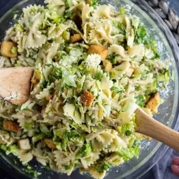 A bowl of Caesar salad pasta salad with farfalle, chopped greens, croutons, and grated cheese is being mixed with wooden utensils for an easy pasta salad perfect for any meal.