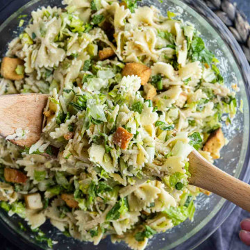 A bowl of Caesar salad pasta salad with farfalle, chopped greens, croutons, and grated cheese is being mixed with wooden utensils for an easy pasta salad perfect for any meal.