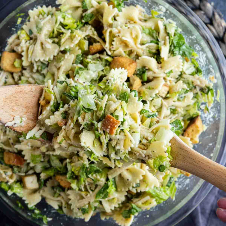 A bowl of Caesar salad pasta salad with farfalle, chopped greens, croutons, and grated cheese is being mixed with wooden utensils for an easy pasta salad perfect for any meal.