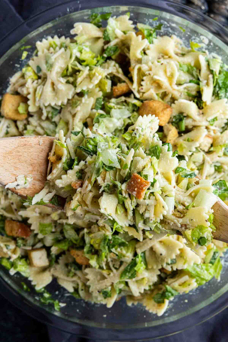 A bowl of bowtie pasta salad mixed with chopped lettuce, croutons, grated cheese, and dressing, with two wooden utensils.