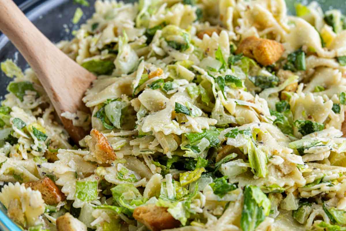 A close-up of a cold pasta salad with bowtie pasta, chopped lettuce, croutons, and a creamy dressing, mixed in a bowl with a wooden spoon—a fresh twist on the classic Caesar salad pasta salad recipe.