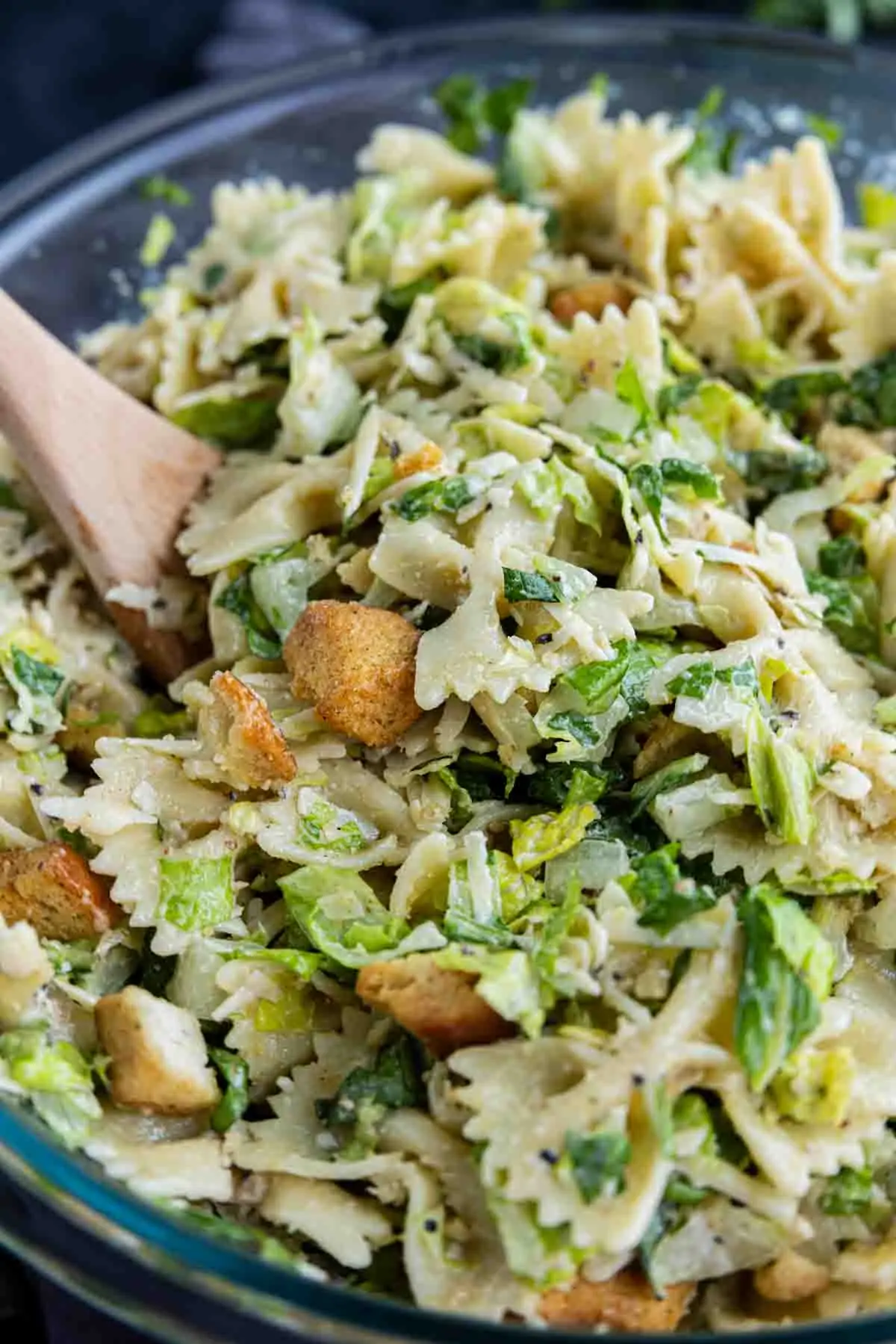 A wooden spoon in a bowl of Caesar salad pasta salad with bowtie pasta, lettuce, croutons, and a creamy dressing—an easy pasta salad perfect for any occasion.