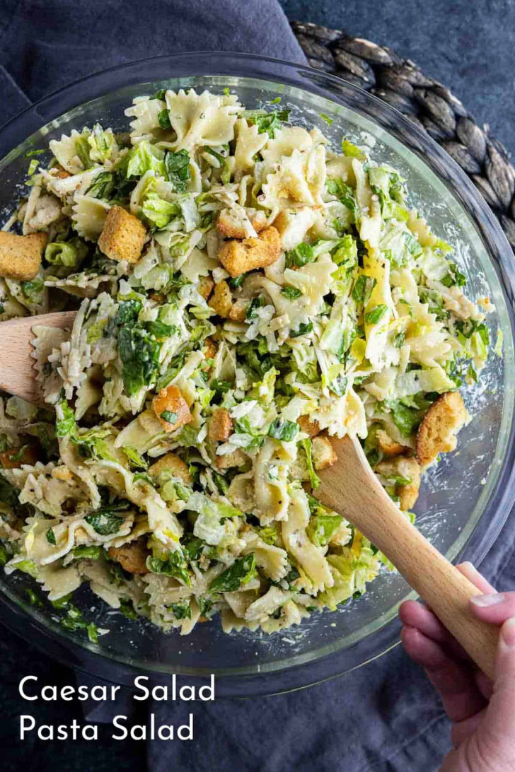 A large glass bowl filled with Caesar salad pasta, featuring bowtie pasta, lettuce, croutons, and grated cheese, being mixed with wooden utensils.
