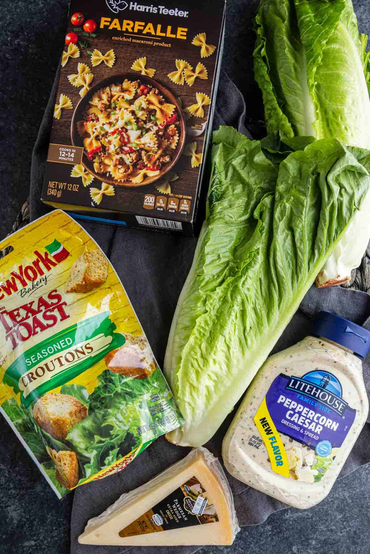 A box of farfalle pasta, romaine lettuce, seasoned croutons, peppercorn Caesar dressing, and Parmesan—everything you need for a delicious Caesar pasta salad recipe on a dark surface.