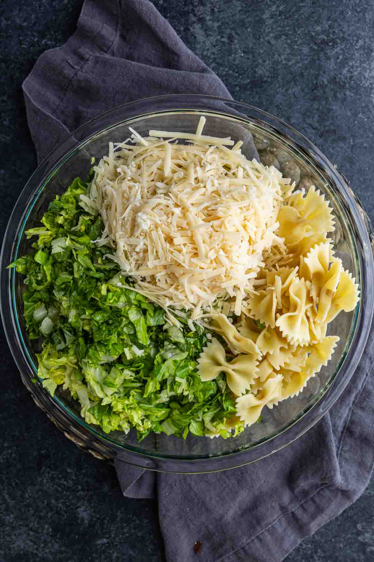 A glass bowl containing a cold pasta salad with cooked bowtie pasta, shredded lettuce, and grated cheese, placed on a dark surface with a gray cloth underneath.