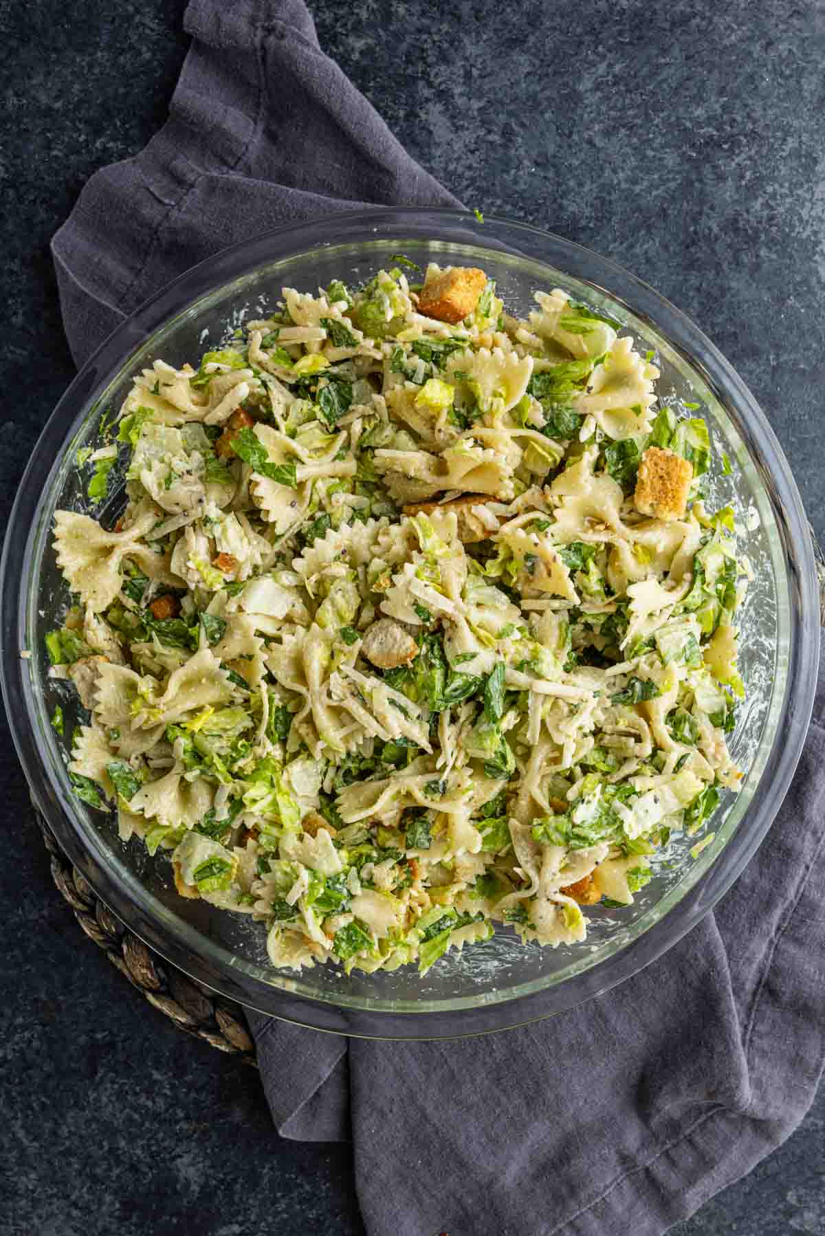 A glass bowl filled with an easy pasta salad of bowtie pasta, chopped lettuce, shredded chicken, croutons, and cheese sits on a dark surface with a gray cloth.