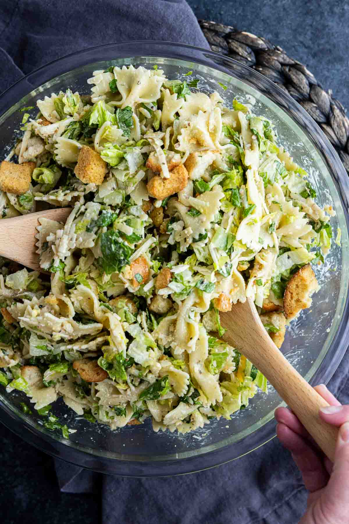 A glass bowl filled with Caesar pasta salad—bowtie pasta mixed with lettuce, croutons, shredded cheese, and creamy dressing—being tossed with wooden utensils for the perfect cold pasta salad.