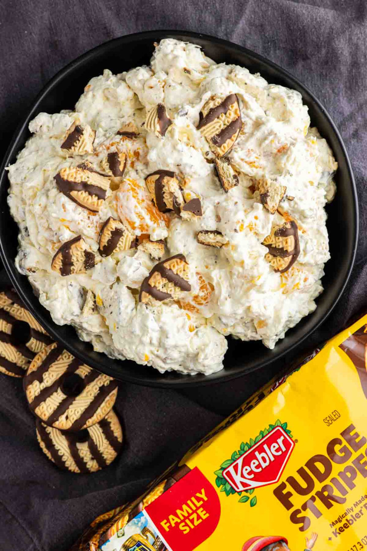 A bowl of creamy Fudge Stripe Cookie Salad topped with pieces of Keebler Fudge Stripes cookies, with whole cookies and a package of Keebler Fudge Stripes beside the bowl—an easy Cookie Salad perfect for any sweet craving.