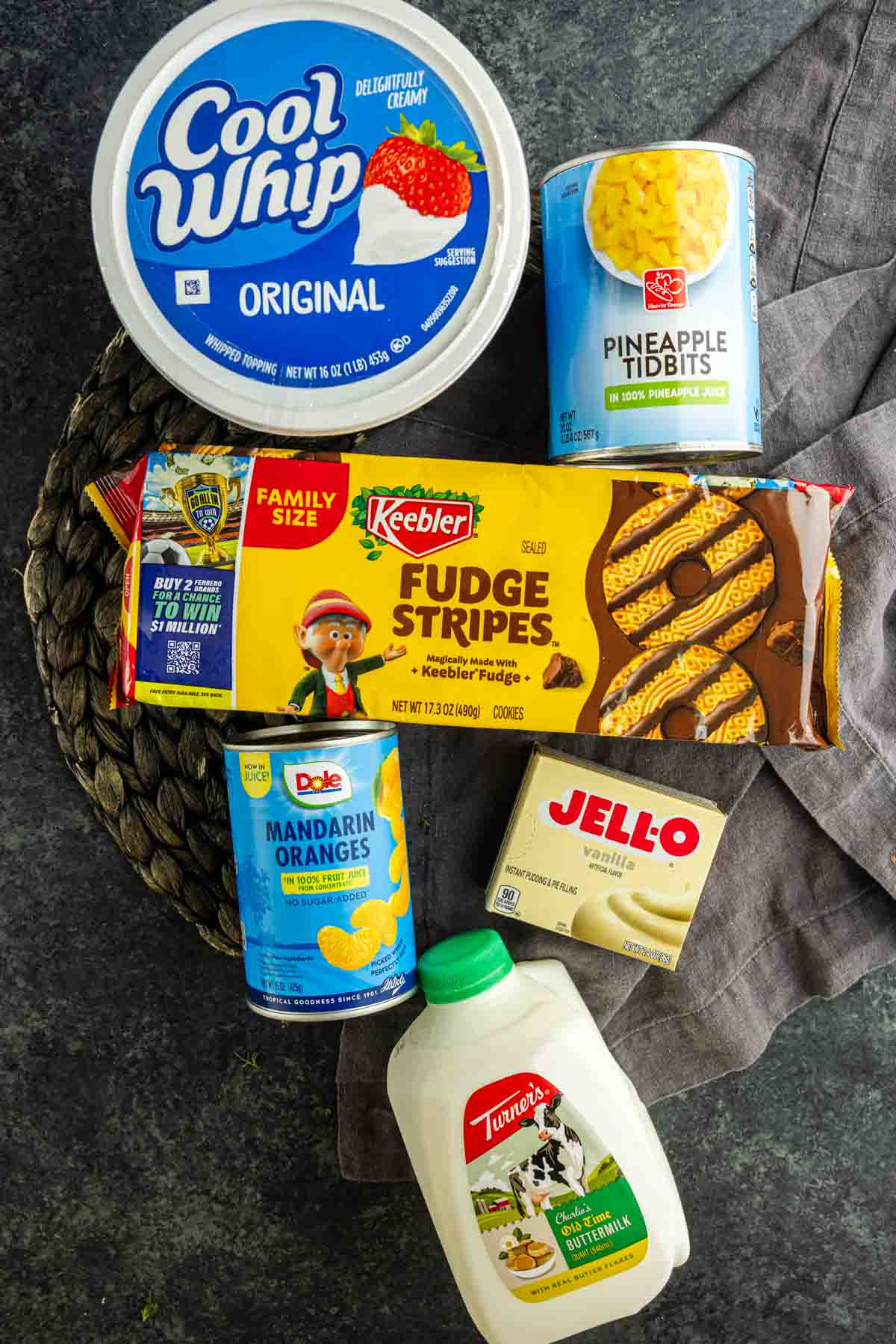 Assorted dessert ingredients for an easy Cookie Salad: Cool Whip, pineapple tidbits, Fudge Stripes cookies, mandarin oranges, vanilla Jello pudding mix, and a carton of milk arranged on a dark surface.