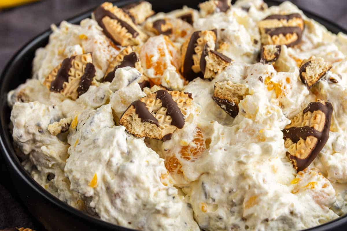 A close-up of a delicious Cookie Salad featuring whipped cream, mandarin orange slices, and pieces of chocolate-covered cookies in a black bowl.