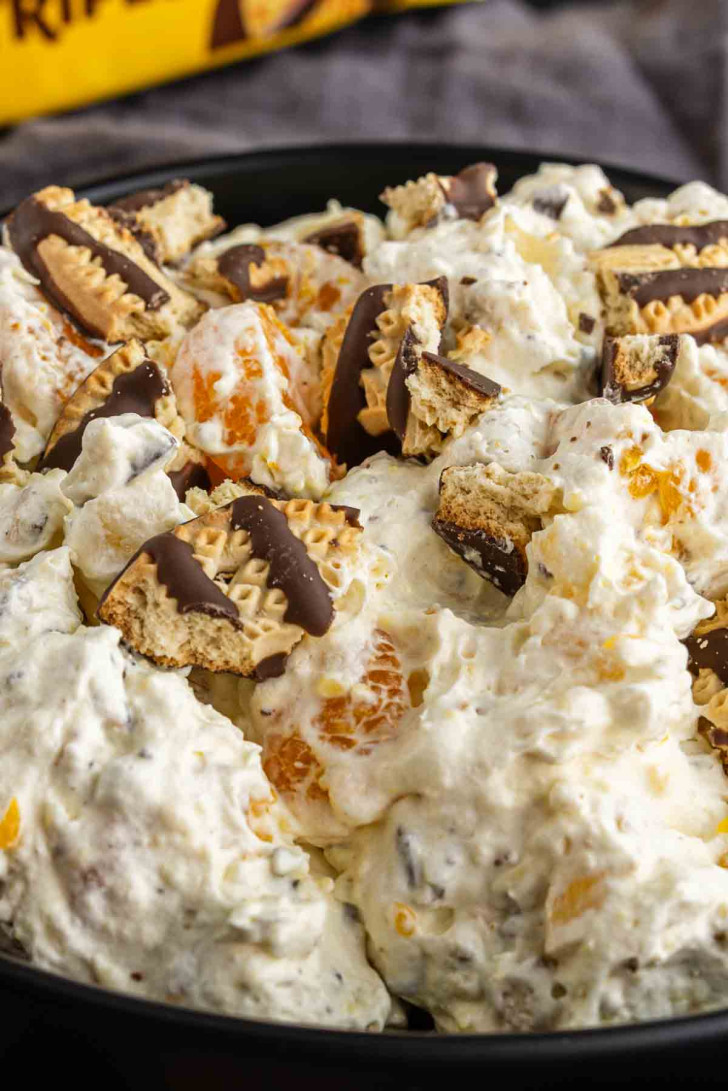 A close-up of a dessert salad with whipped cream, mandarin orange segments, mini marshmallows, and chopped chocolate-dipped cookies in a black bowl.