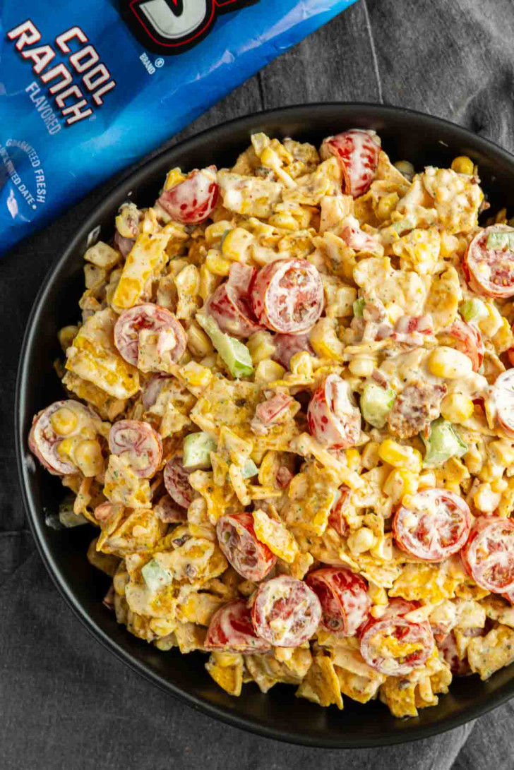 A black bowl filled with salad made of cherry tomatoes, corn, chopped vegetables, and crumbled Doritos, with a blue bag of Cool Ranch Doritos beside it.