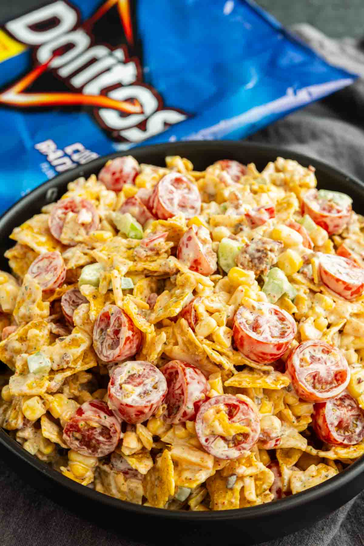 A bowl of Cool Ranch Doritos Salad with chopped tomatoes, corn, green onions, and creamy dressing, with a bag of Doritos chips in the background.