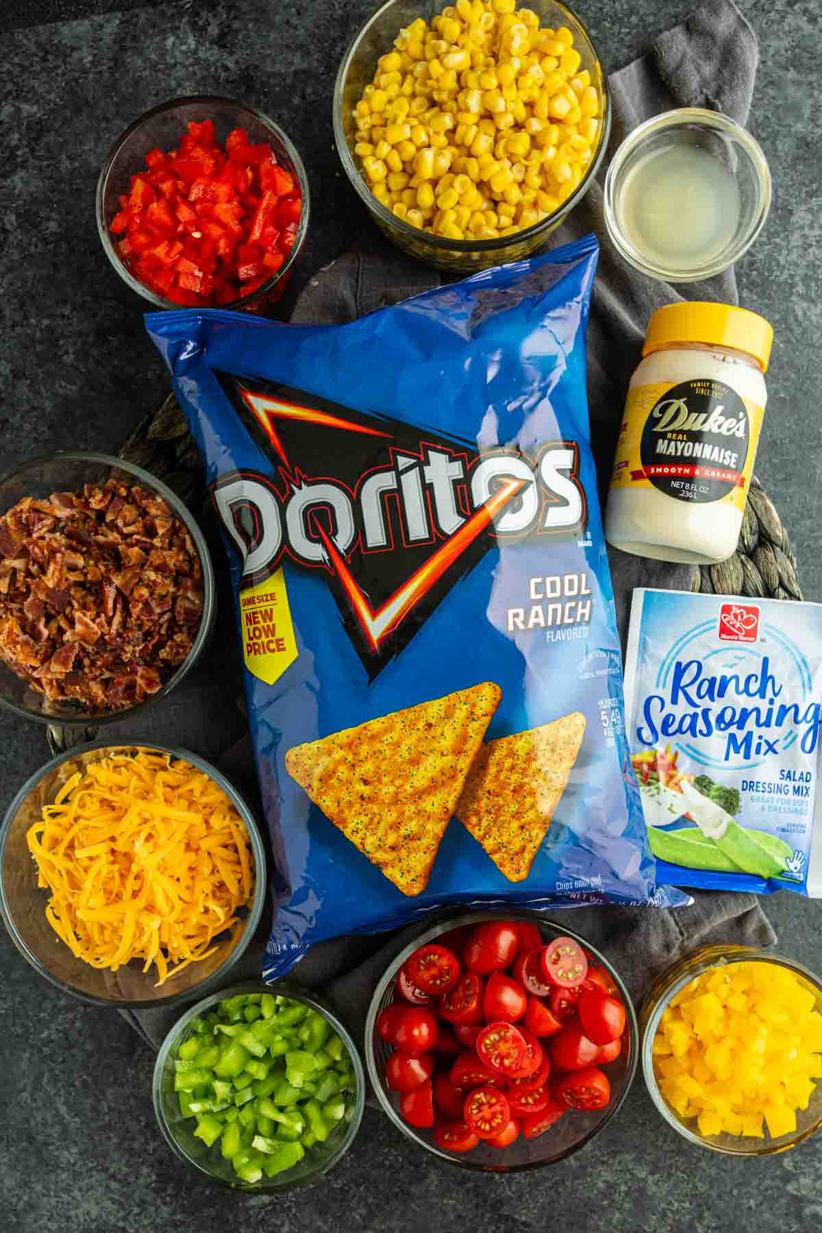 A bag of Cool Ranch Doritos is surrounded by bowls of chopped red pepper, corn, bacon, cheddar cheese, celery, tomatoes, green pepper, mayonnaise, and a packet of ranch seasoning—everything you need for a flavorful Cool Ranch Doritos Salad.