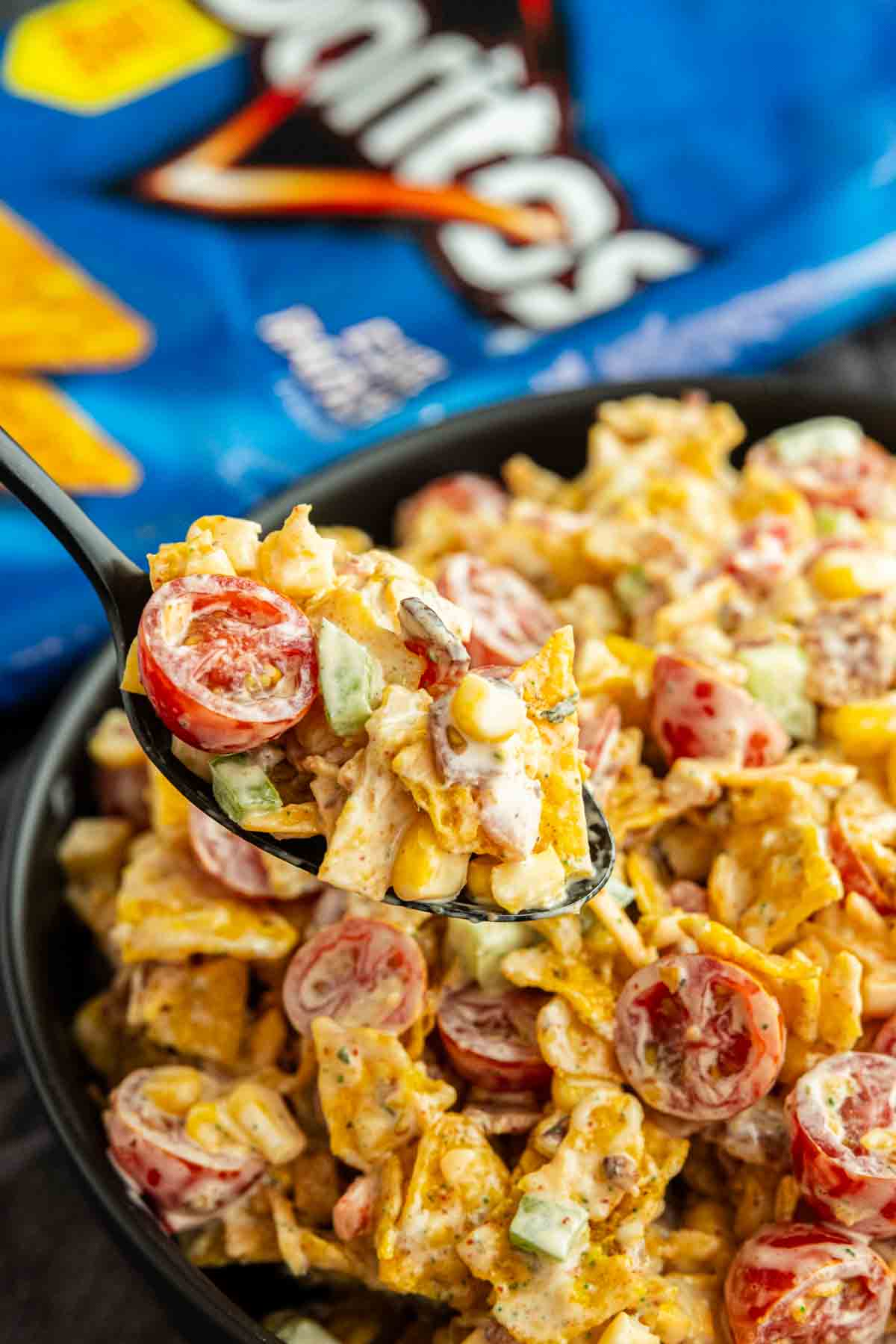 A spoonful of Cool Ranch Doritos Salad with cherry tomatoes, corn, cucumber, and creamy dressing, with a bag of Doritos in the background.