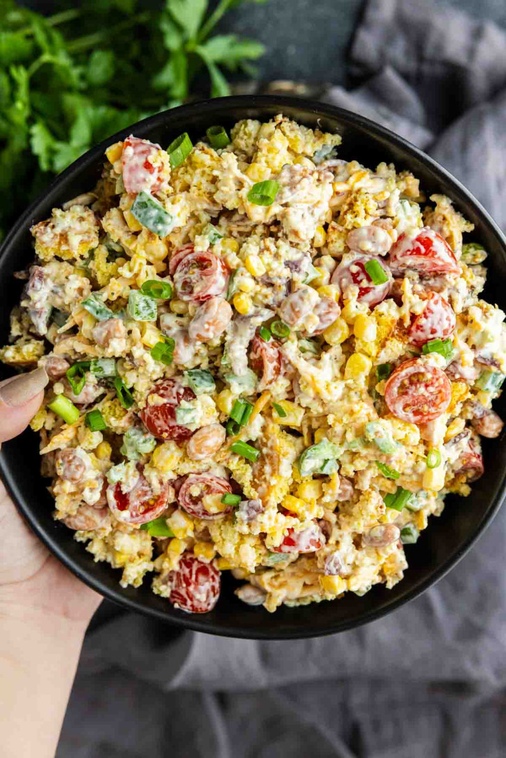 A bowl of salad with cherry tomatoes, corn, chopped green onions, and creamy dressing is held by a hand—perfect as a Southern potluck side dish. Fresh parsley adds color in the background, making it ideal for any summer BBQ side.