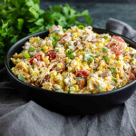 A black bowl filled with Southern Cornbread Salad—mixed greens, cherry tomatoes, corn, green onions, and shredded cheese—set on a gray cloth with fresh herbs in the background; a perfect Southern potluck side dish.