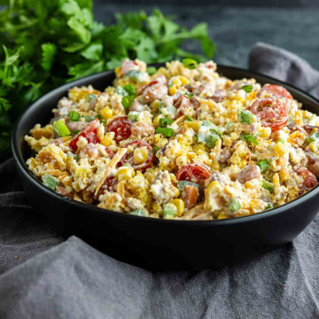 A black bowl filled with Southern Cornbread Salad—mixed greens, cherry tomatoes, corn, green onions, and shredded cheese—set on a gray cloth with fresh herbs in the background; a perfect Southern potluck side dish.
