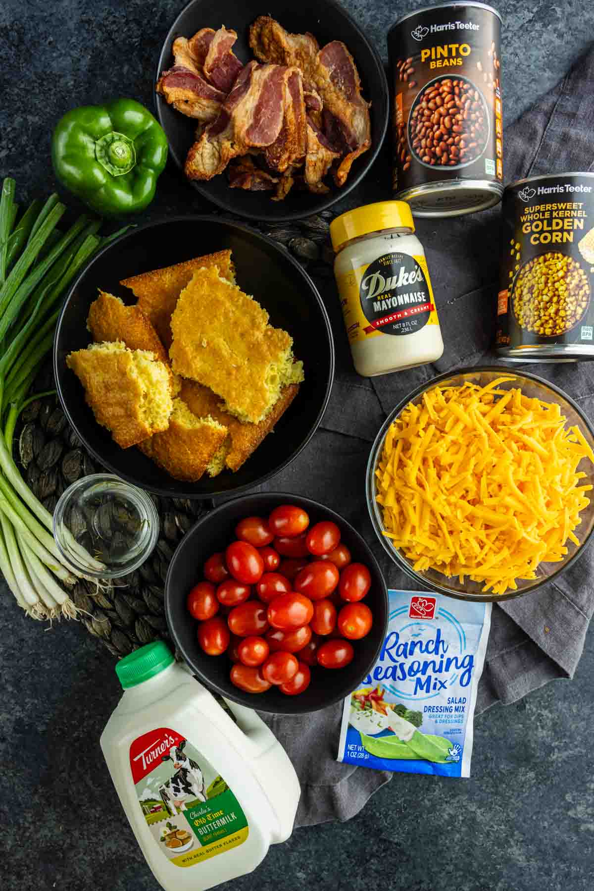 Ingredients for a cornbread salad with ranch dressing arranged on a dark surface, perfect as a summer BBQ side—cornbread, bacon, cheddar, grape tomatoes, ranch mix, mayo, pinto beans, corn, green onions, green pepper, and buttermilk.