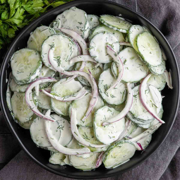 A black bowl filled with creamy cucumber salad, featuring sliced cucumbers, red onions, fresh dill, and a rich sour cream dressing.