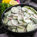 A black bowl filled with creamy cucumber salad made of cucumber and red onion slices in a dill dressing, with parsley and lemon in the background.
