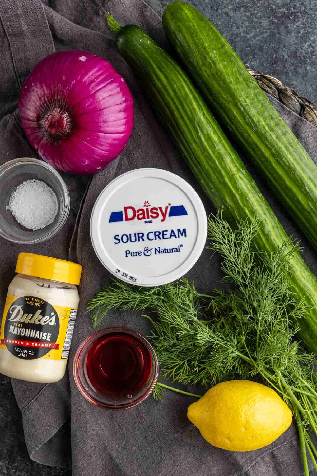 Top-down view of ingredients for creamy cucumber salad on a cloth: red onion, cucumbers, fresh dill, lemon, sour cream, mayonnaise, salt, and a small bowl of red liquid.