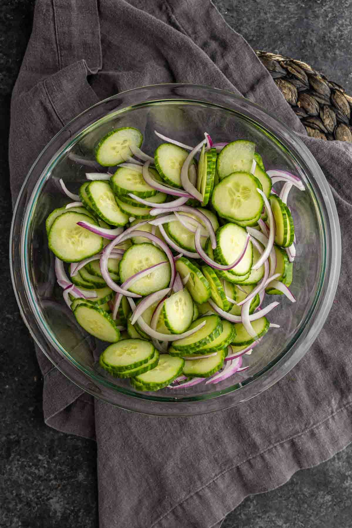 A glass bowl filled with sliced cucumbers and thinly sliced red onions, ready for a refreshing cucumber onion salad, sits on a dark cloth and countertop.