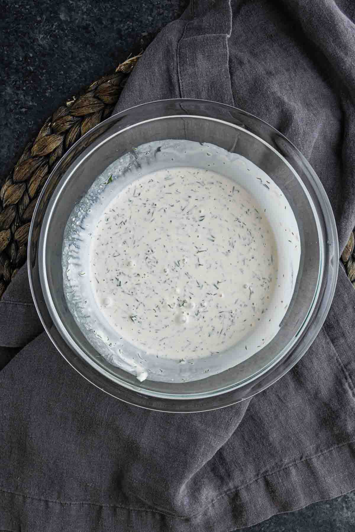 A glass bowl filled with creamy ranch dressing, perfect for a dill cucumber salad, sits on a dark gray cloth and woven mat.