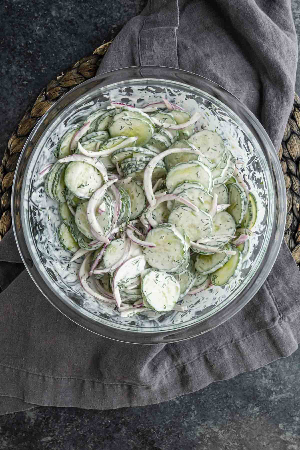 A glass bowl filled with creamy cucumber salad and red onions, garnished with dill, sits on a gray cloth and woven mat.