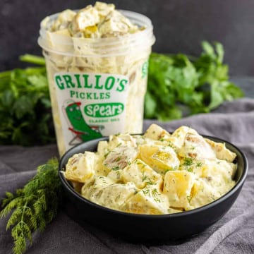 A bowl of creamy dill pickle potato salad, garnished with dill, sits in front of a container of Grillo’s Pickles spears, with fresh herbs in the background.